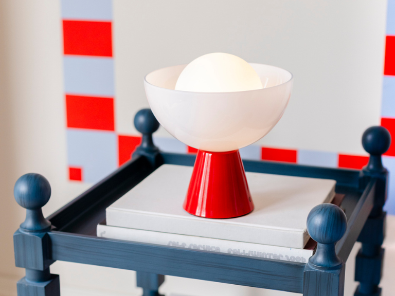 A portable, wireless lamp with a red-glossy metal base sits on a navy blue bedside table on top of a few books. The lamp features a white milky glass bowl that encases an opal glass shade with an integrated, rechargeable LED bulb.