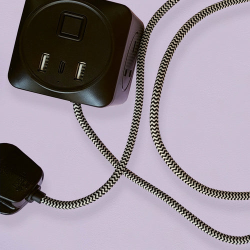 A black cuboid extension lead with three plug sockets and three USB sockets, including one USB-C port with black and white herringbone fabric on a lilac background.