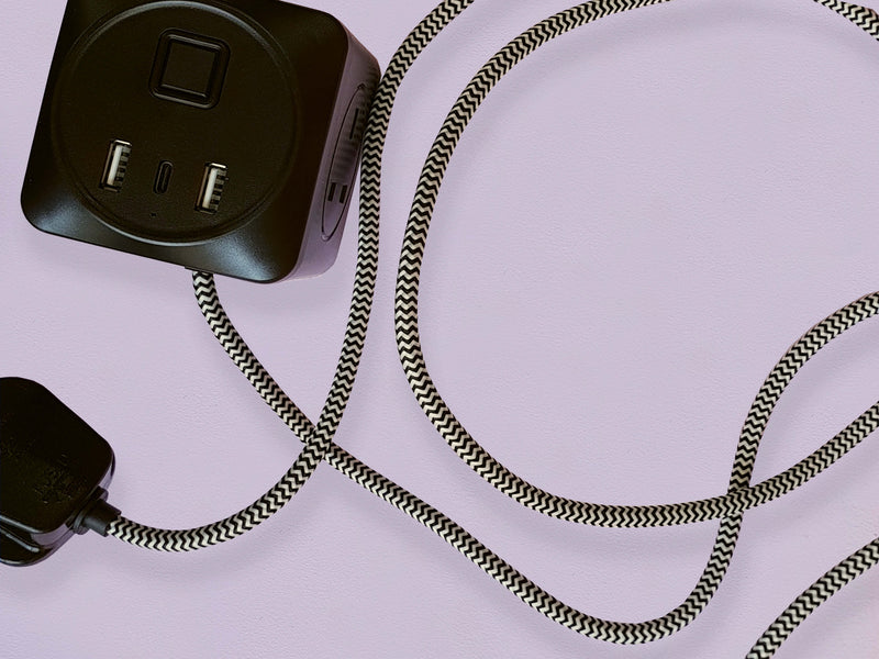 A black cuboid extension lead with three plug sockets and three USB sockets, including one USB-C port with black and white herringbone fabric on a lilac background.