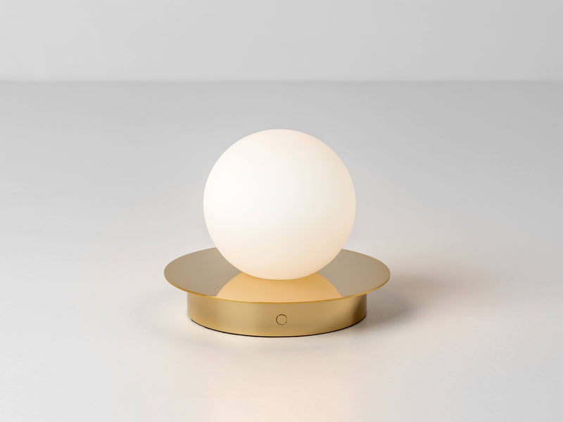 An opal glass shade sits upon a metal disk. The opal glass shade contains an integrated LED bulb that can be recharged. The lamp is portable and wireless. Light is on.