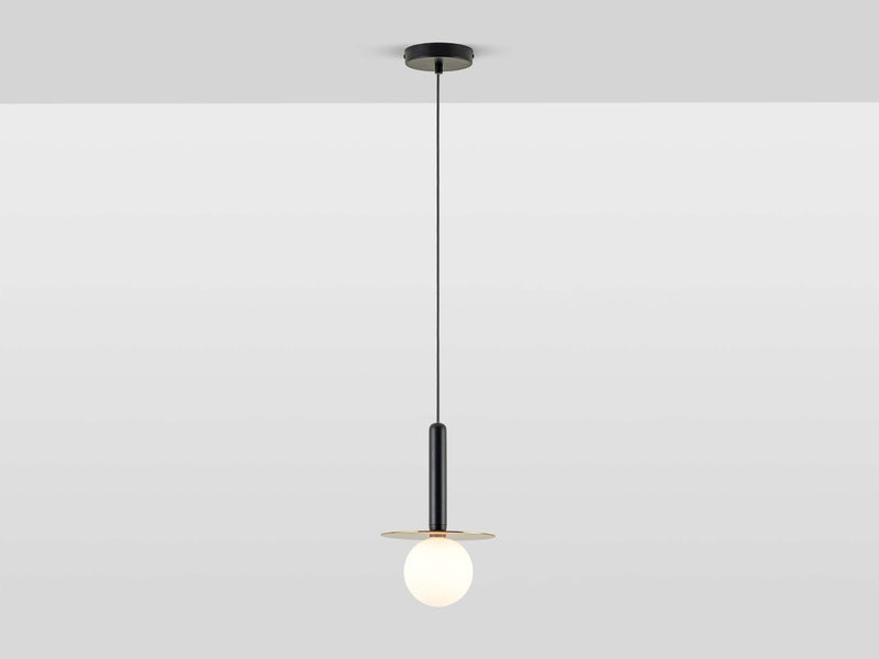 A pendant ceiling light with a charcoal grey oval body oval body and a horizontal brass metal plate that holds a white opal glass shade. The cord is black and the light is switched on.