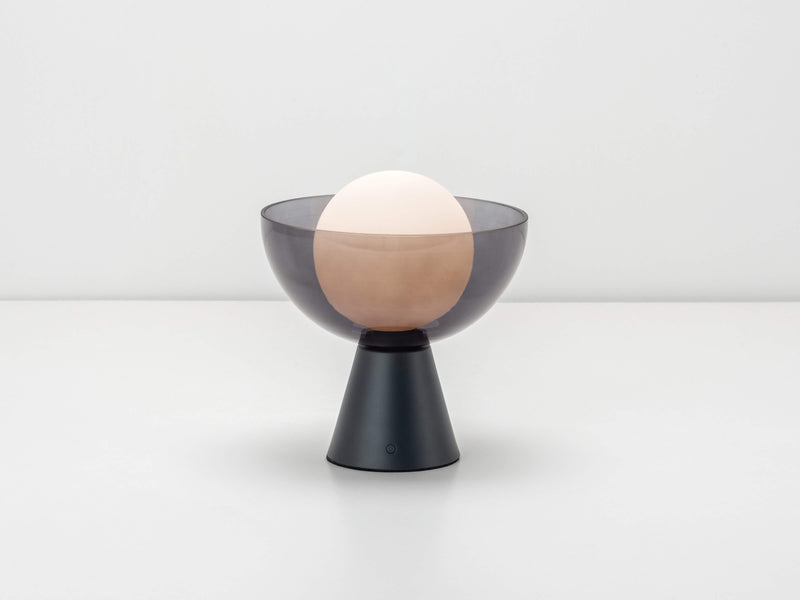 Upon a metal base sits a charcoal smoked glass bowl, that encases an opal glass shade. The glass shade contains an integrated LED bulb that can be recharged. 