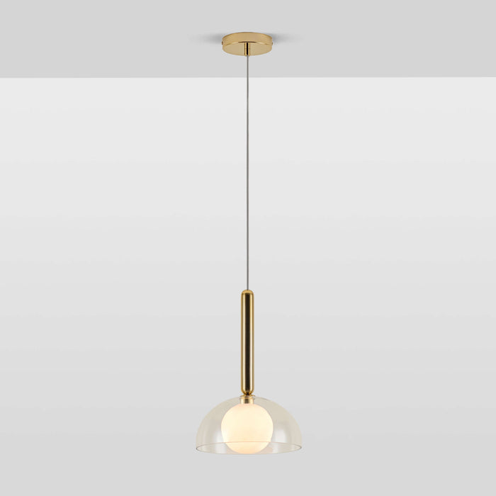 A pendant light that features a brass oval metal body attached to a clear glass dome shade which encases an opal glass shade. The light is switched on.