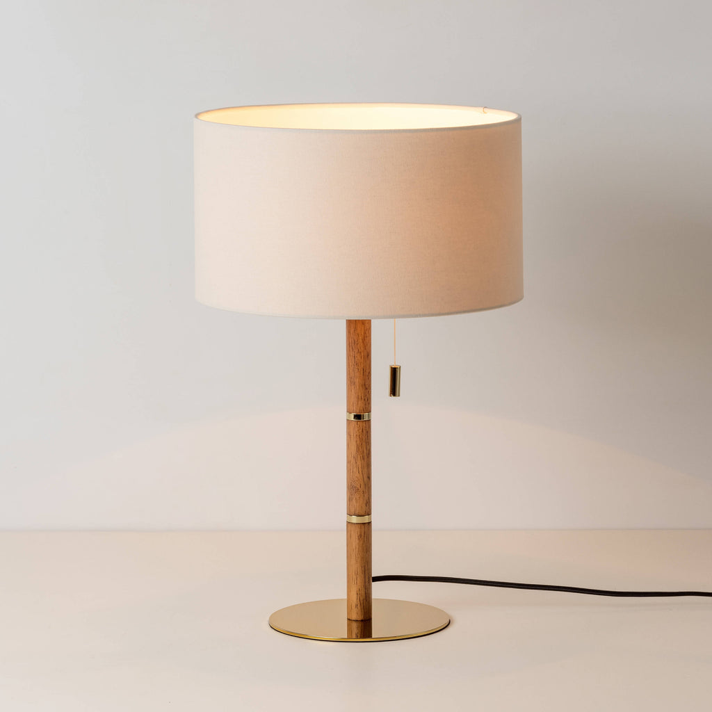 A linen drum shade sits upon a wooden stick base, with equidistant brass counters inserted into the wooden base and matching brass base, with brass pull switch. Light is on.