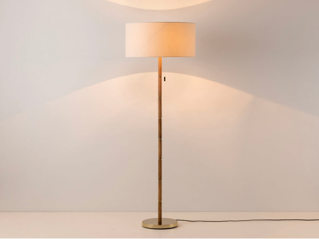 A large linen drum shade sits upon a wooden stick base, with equidistant brass counters inserted into the wooden base and matching brass base. The light is on.